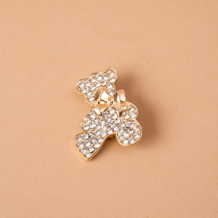 The Crystal Teddy Bear Timeless Brooch by The Darling Effect is a gold-tone teddy bear brooch covered in clear crystals with a small bow at the neck, serving as a vintage-inspired keepsake accessory.
