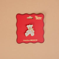 The Crystal Teddy Bear Timeless Brooch by The Darling Effect is a gold-toned, vintage-inspired teddy bear pin encrusted with rhinestones and wearing a small bow, attached to a red display card as a timeless keepsake accessory.