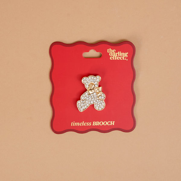 The Crystal Teddy Bear Timeless Brooch by The Darling Effect is a gold-toned, vintage-inspired teddy bear pin encrusted with rhinestones and wearing a small bow, attached to a red display card as a timeless keepsake accessory.