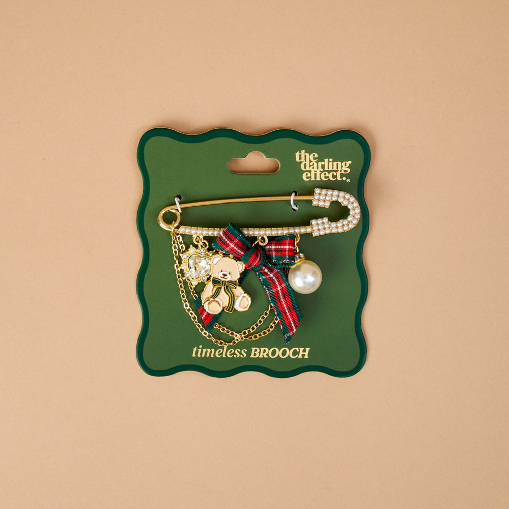 A Holiday Heritage Timeless Brooch by The Darling Effect featuring a vintage-inspired gold safety pin with a teddy bear charm, plaid ribbon bow, pearl, and crystal heart, mounted on a green display card.