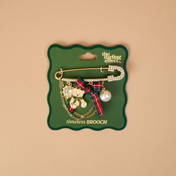 A Holiday Heritage Timeless Brooch by The Darling Effect featuring a vintage-inspired gold safety pin with a teddy bear charm, plaid ribbon bow, pearl, and crystal heart, mounted on a green display card.