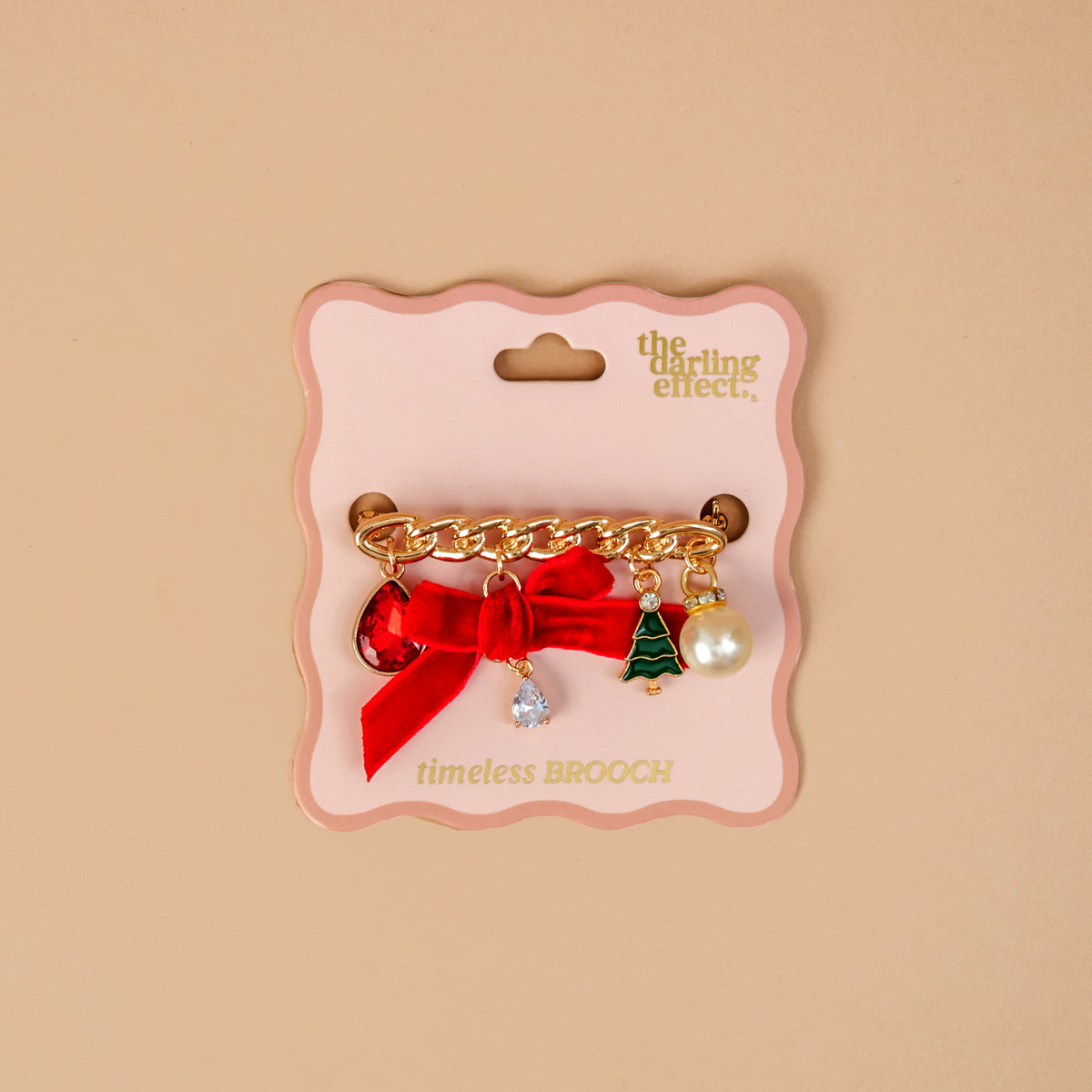 Holiday Sparkle Timeless Brooch by The Darling Effect, a vintage-inspired gold-tone chain brooch with a red ribbon bow, red and clear teardrop charms, a green Christmas tree charm, and a faux pearl, mounted on a scalloped display card.