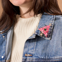 A Whimsical Tree Timeless Brooch, a vintage-inspired pink Christmas tree with colorful accents, pinned to the collar of a denim jacket.
