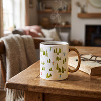 An Aprés Ski 11 oz Cozy Cute Ceramic Mug with a pattern of evergreen trees and skiers, a gold handle, and rising steam sits on a wooden table next to an old book and glasses for a cozy moment.