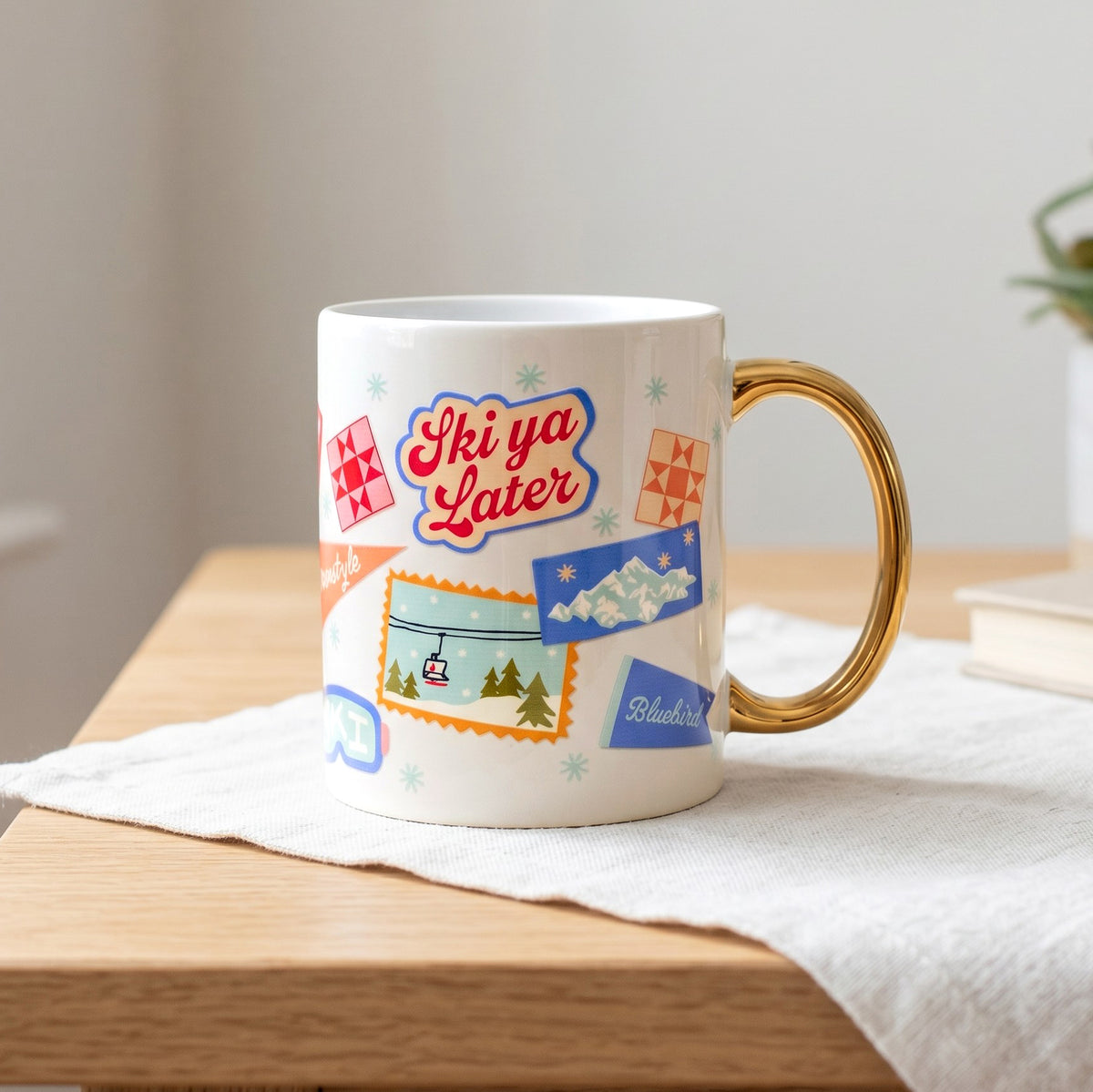 A cozy white Ski Ya Later 11 oz Cozy Cute Ceramic Mug by The Darling Effect featuring a gold handle and ski-themed stickers of a mountain scene, a ski lift, and the text Ski ya Later.