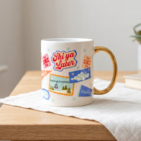 A cozy white Ski Ya Later 11 oz Cozy Cute Ceramic Mug by The Darling Effect featuring a gold handle and ski-themed stickers of a mountain scene, a ski lift, and the text Ski ya Later.