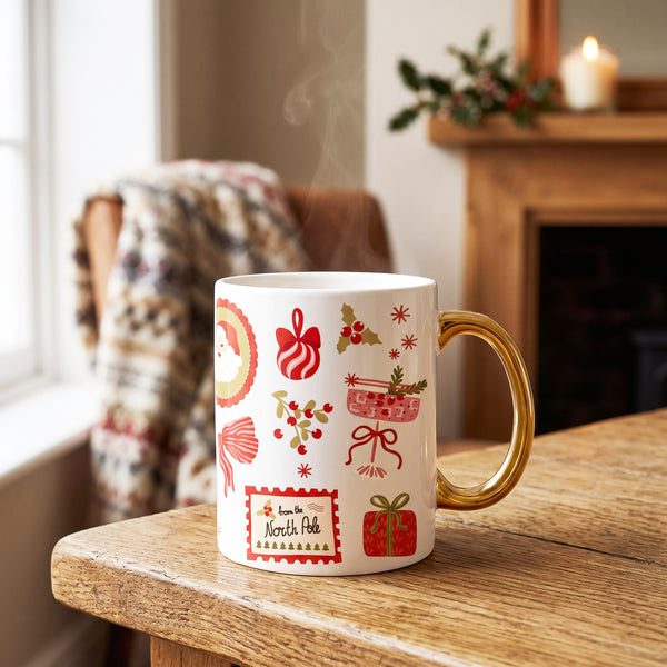 A Mistletoe Magic 11 oz Cozy Cute Ceramic Mug by The Darling Effect with a gold handle and Christmas illustrations sits on a wooden table, steam rising from the North Pole-labeled cup.