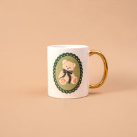 Teddy Bear Theo 11 oz Cozy Cute Ceramic Mug in white with a gold handle, featuring a green scalloped oval emblem of a teddy bear wearing a dark plaid ribbon.