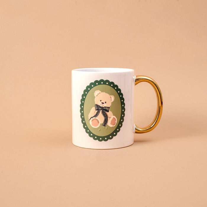 Teddy Bear Theo 11 oz Cozy Cute Ceramic Mug in white with a gold handle, featuring a green scalloped oval emblem of a teddy bear wearing a dark plaid ribbon.