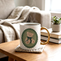 Teddy Bear Theo 11 oz Cozy Cute Ceramic Mug: white ceramic with a gold handle, featuring a graphic of a teddy bear wearing a green plaid bow inside a scalloped oval frame.