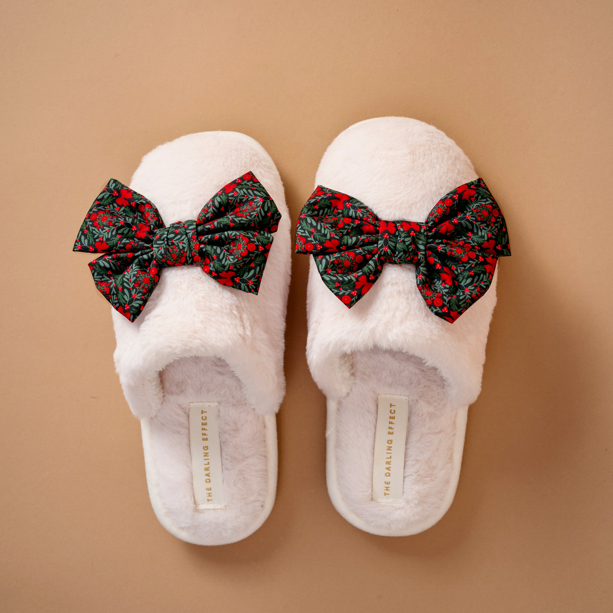 Betty Bow A Merry Moment On Cloud Nine Slippers in cream-colored faux fur, each topped with a large bow in a festive green and red holiday floral print.