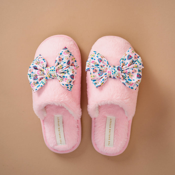 Betty Bow Sugarplum Lane On Cloud Nine Slippers, a pair of pink faux fur slippers, each featuring a large bow with a colorful pattern of houses and trees.
