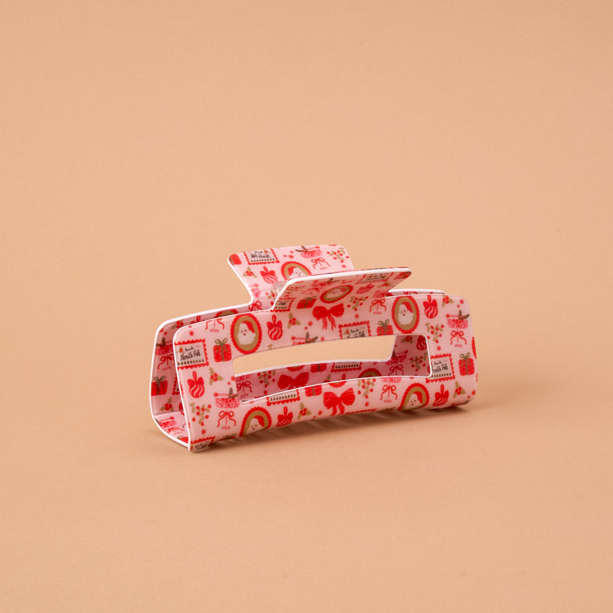 A Mistletoe Magic Claw Clip featuring a rectangular design with a repeating pattern of red bows, gift boxes, and festive wreaths on a light pink background.