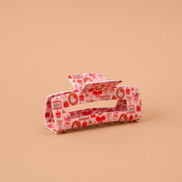 A Mistletoe Magic Claw Clip featuring a rectangular design with a repeating pattern of red bows, gift boxes, and festive wreaths on a light pink background.
