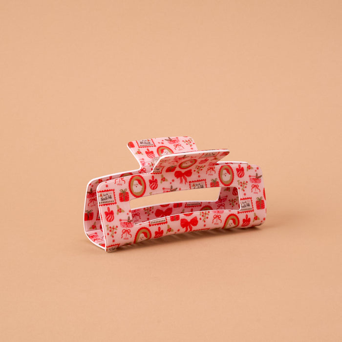 A Mistletoe Magic Claw Clip featuring a rectangular design with a repeating pattern of red bows, gift boxes, and festive wreaths on a light pink background.