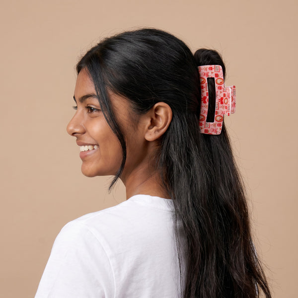 A person seen from the side wearing a pink printed Mistletoe Magic Claw Clip by The Darling Effect, decorated with small holiday-themed icons.