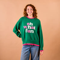 A woman wears The Darling Effect Oh What Fun Classic Sweater, a festive green knit with pink trim and oh what fun in textured pink and white lettering across the chest.