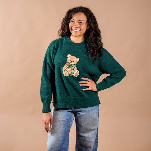 A woman smiling while wearing The Darling Effect Teddy Bear Theo Classic Sweater, which features a festive teddy bear appliqué with a green bow.