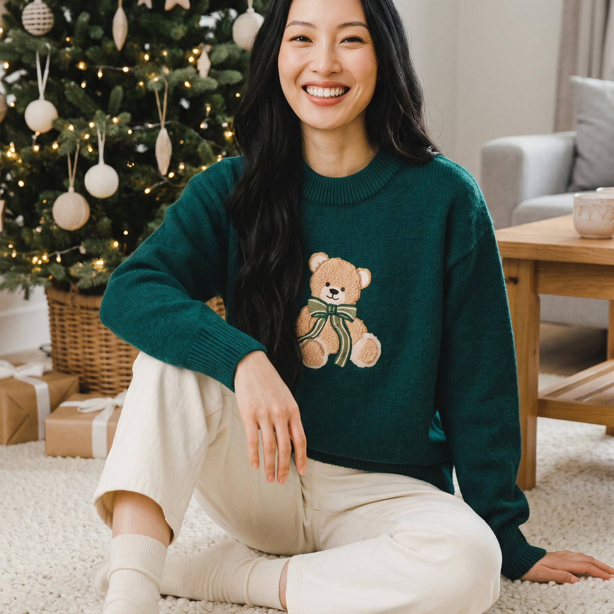 A smiling woman wears a dark green Teddy Bear Theo Classic Sweater featuring an embroidered teddy bear with a green ribbon while sitting on the floor in front of a Christmas tree.