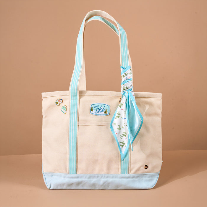 Aprés Ski The Darling Canvas Tote Bag in cream canvas with light blue striped straps, a blue Après Ski patch, two small ski-themed pins, and a patterned neckerchief tied to the handle.