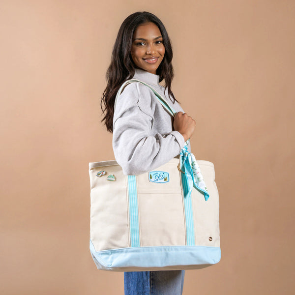 A person smiles while carrying an Aprés Ski The Darling Canvas Tote Bag, featuring a spacious design with a light blue bottom, striped handles, and an embroidered Aprés Ski patch.