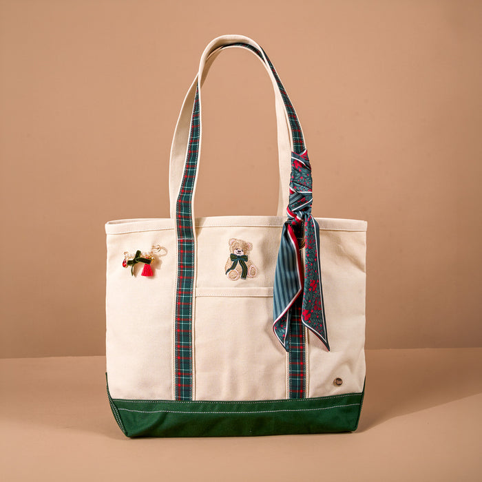 Evergreen Plaid The Darling Canvas Tote Bag in off-white canvas with a dark green bottom, plaid strap trim, an embroidered teddy bear, a holiday pin, and a patterned scarf, perfect as a work travel bag.