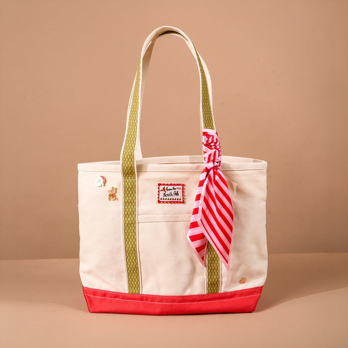 The Darling Effect Peppermint Stripes The Darling Canvas Tote Bag is an off-white canvas tote with olive green striped handles, a red base, a From the North Pole patch, two pins, and a red-and-white striped ribbon tied to one handle.
