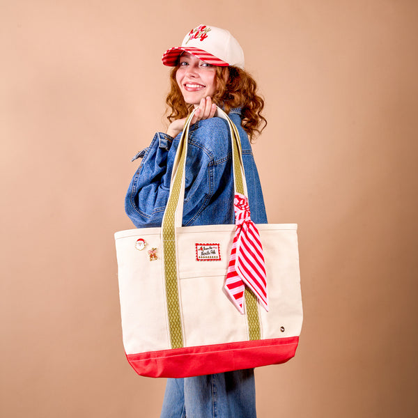 A person in a denim jacket holds a Peppermint Stripes The Darling Canvas Tote Bag, featuring red trim, green webbing handles, a red-and-white striped ribbon, and a North Pole patch.