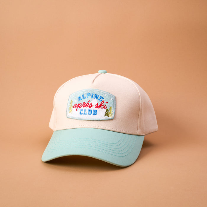 The Aprés Ski Club Classic Canvas Hat is a light pink casual hat with a light blue brim and a rectangular embroidered patch that reads Alpine après ski CLUB with small pine tree icons.