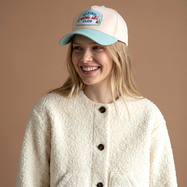 The Aprés Ski Club Classic Canvas Hat is a cream baseball cap with a light blue brim and an Alpine après ski club patch on the front.