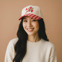 A woman smiles wearing a cream-colored Holly Jolly Classic Canvas Hat with a red-and-white striped brim and holly jolly embroidered in red and gold on the front.