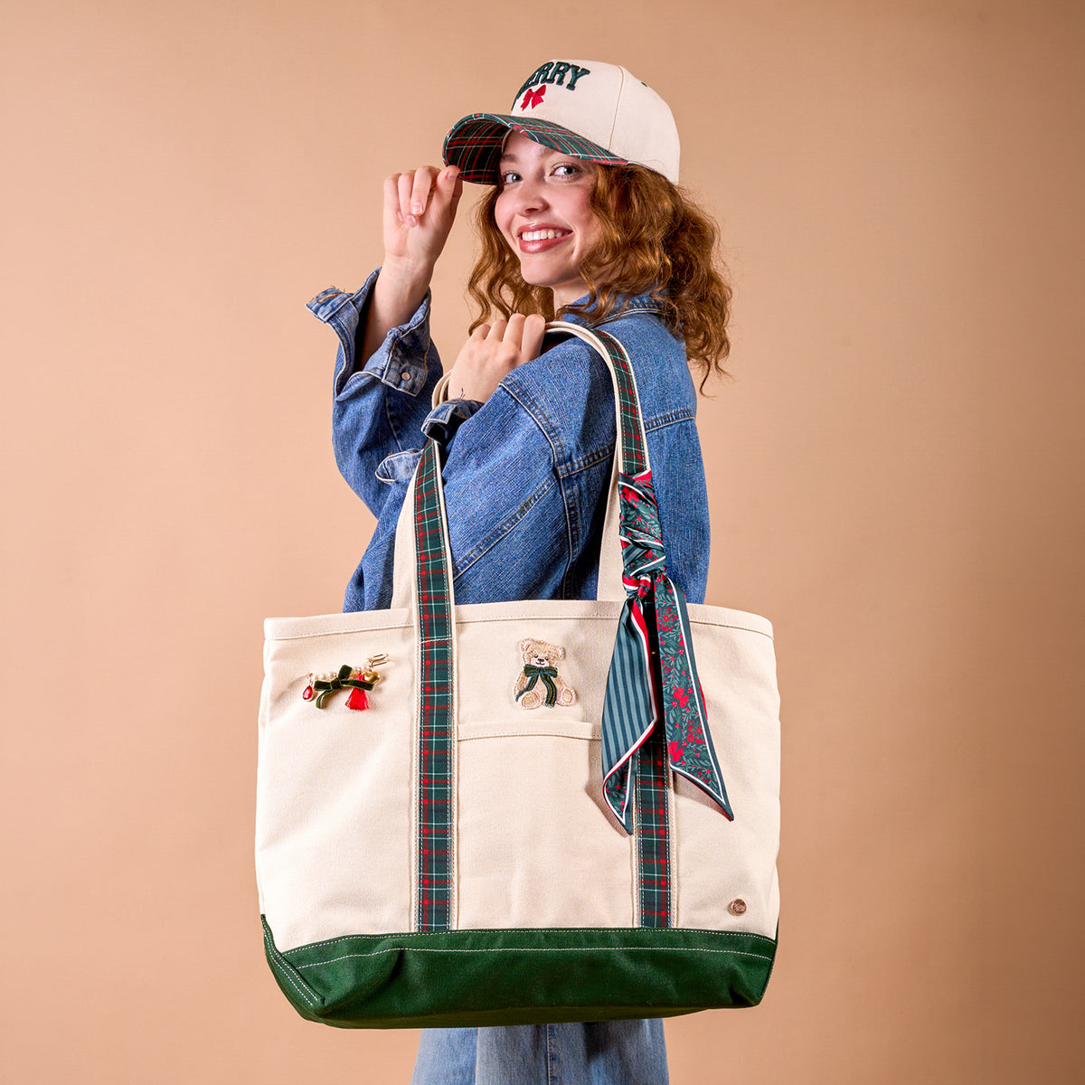 A woman in a denim jacket and a Merry Classic Canvas Hat holds a large canvas tote bag decorated with ribbon handles, a bear patch, and a festive pin.