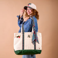 A woman in a denim jacket and a Merry Classic Canvas Hat holds a large canvas tote bag decorated with ribbon handles, a bear patch, and a festive pin.