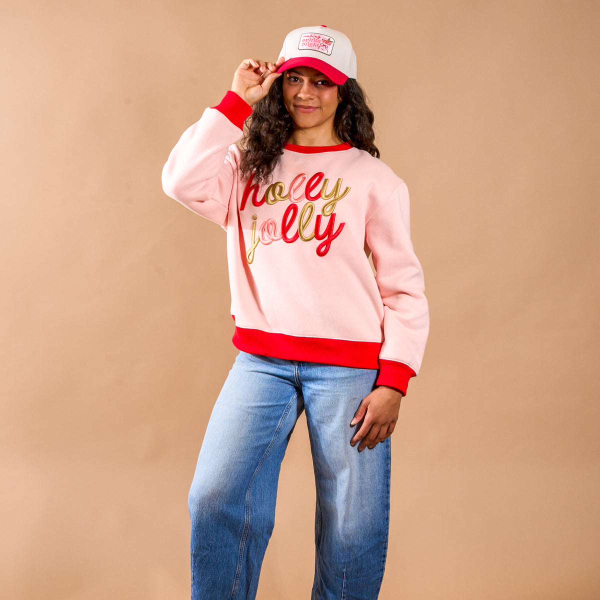 A person wearing a white and red Making Spirits Bright Classic Canvas Hat and a pink sweatshirt with the words holly jolly in gold script across the front.
