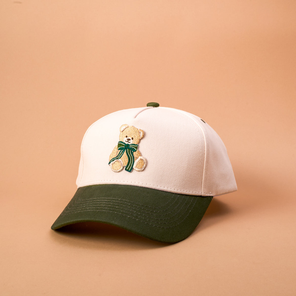 The Darling Effect Teddy Bear Theo Classic Canvas Hat is a cream relaxed-fit baseball cap featuring a dark green brim and an embroidered teddy bear wearing a green ribbon on the front.