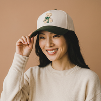 A smiling woman wearing a cream-colored Teddy Bear Theo Classic Canvas Hat with a dark green brim and an embroidered teddy bear wearing a green bow on the front.