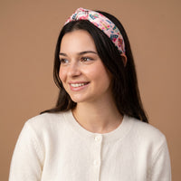 A person wearing a Sugarplum Lane Classic Twist Headband, a pink satin knotted headband patterned with colorful houses.