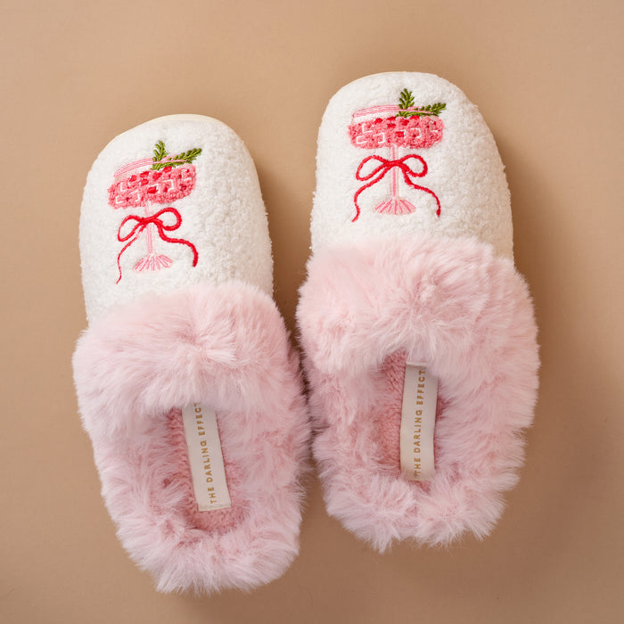 A pair of white, fuzzy Cranberry Cosmo Dreamy Daze slippers by The Darling Effect, featuring pink faux fur trim and an embroidered cocktail glass design on the toe.
