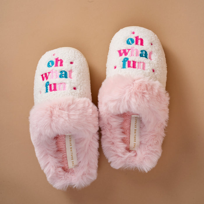 Cream-colored Oh What Fun Dreamy Daze slippers featuring pink faux-fur trim and the embroidered text oh what fun on the top.