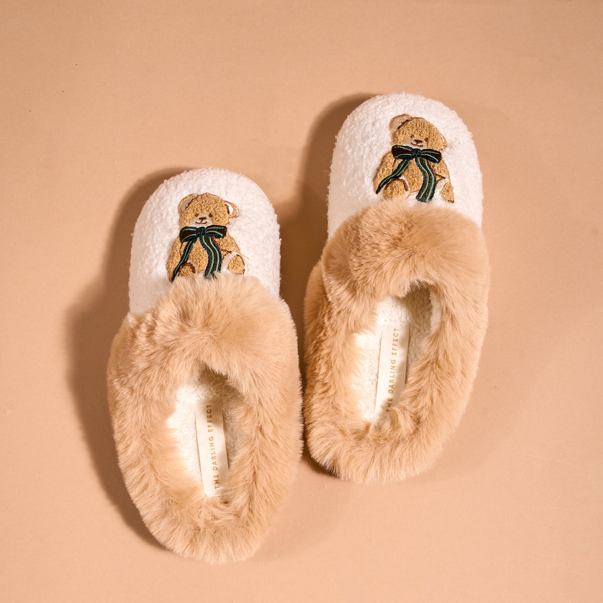 Pair of cream-colored Teddy Bear Theo Dreamy Daze Slippers by The Darling Effect, each featuring an embroidered teddy bear wearing a green ribbon and a cozy faux-fur collar.