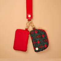 A Merry Moment Dynamic Duo Pouch Wristlet featuring two small zippered pouches hanging from a red strap, one solid red and the other a festive pattern of bows and greenery.
