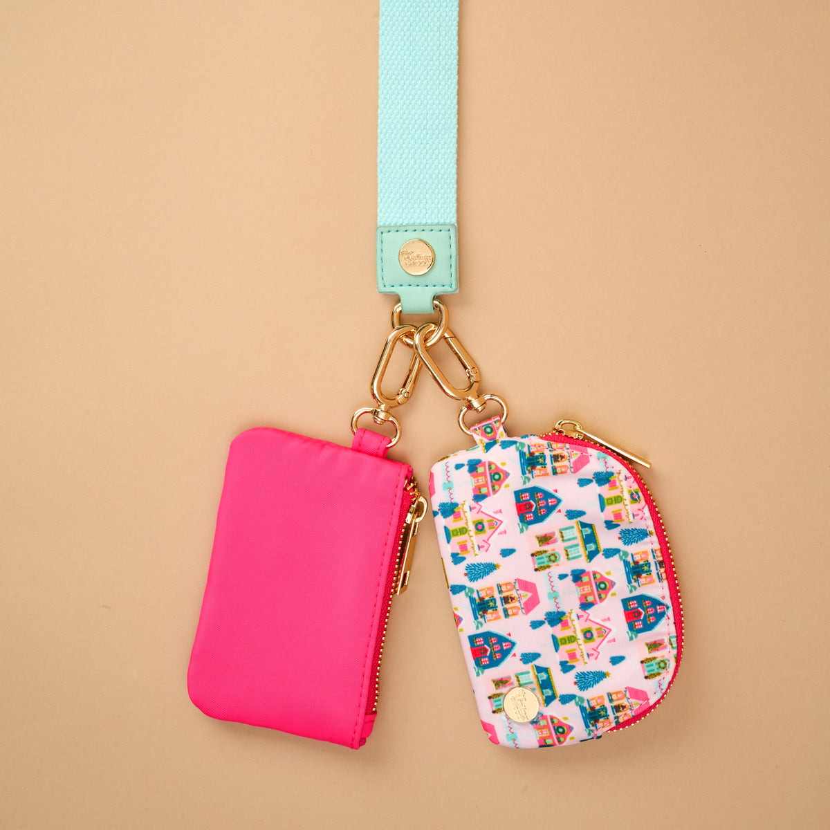 The Sugarplum Lane Dynamic Duo Pouch Wristlet features a neon pink pouch and a holiday village print pouch hanging from a light blue strap with gold-tone hardware, offering a hands-free way to carry your zippered pouches.