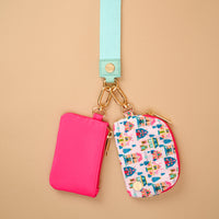 The Sugarplum Lane Dynamic Duo Pouch Wristlet features a neon pink pouch and a holiday village print pouch hanging from a light blue strap with gold-tone hardware, offering a hands-free way to carry your zippered pouches.