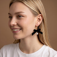 A person wearing Midnight Velvet Signature Hoop Earrings, which are lightweight gold hoops featuring a black ribbon bow and a hanging white pearl.