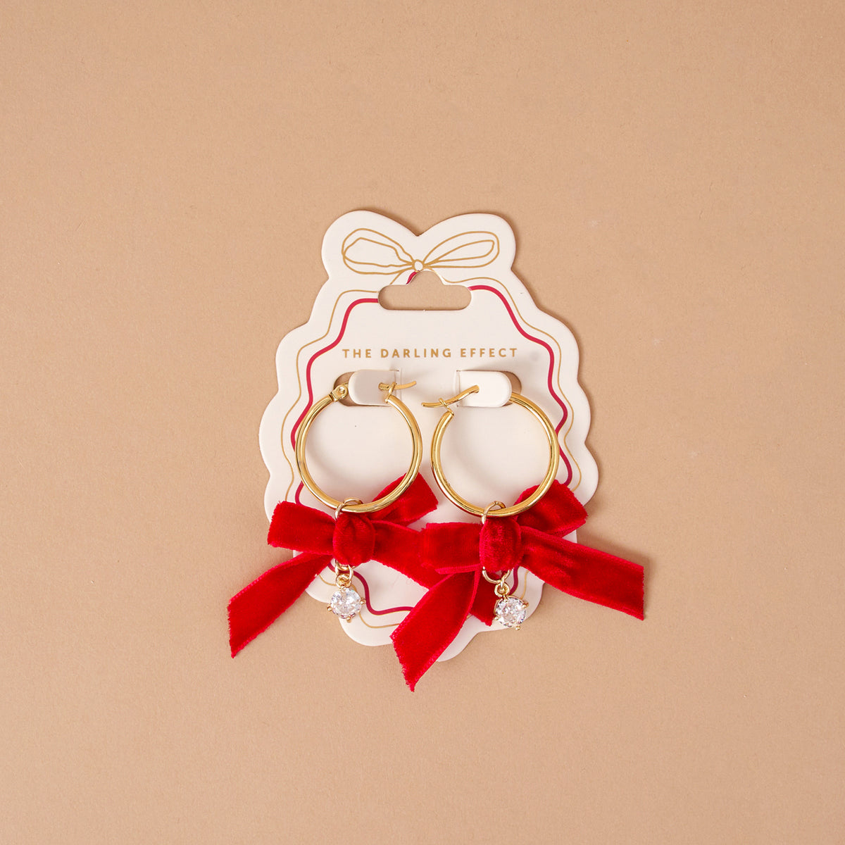 Ruby Ribbon Signature Hoop Earrings from The Darling Effect: lightweight gold hoops featuring red velvet bows and small dangling clear crystals, displayed on a branded white card.