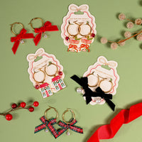 Five pairs of Ruby Ribbon Signature Hoop Earrings by The Darling Effect on display cards, featuring holiday accents including red bows, cocktail glasses, houses, black bows with pearls, and plaid bows.