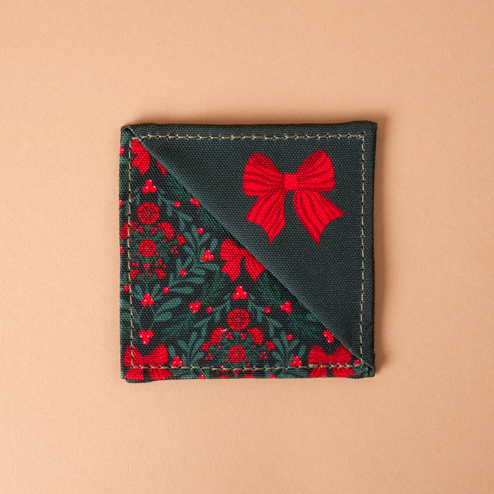A Merry Moment Corner Keeper fabric bookmark, shaped as a triangle with dark green holly leaves and red berries on one side and a solid dark green section with a red bow print on the other.