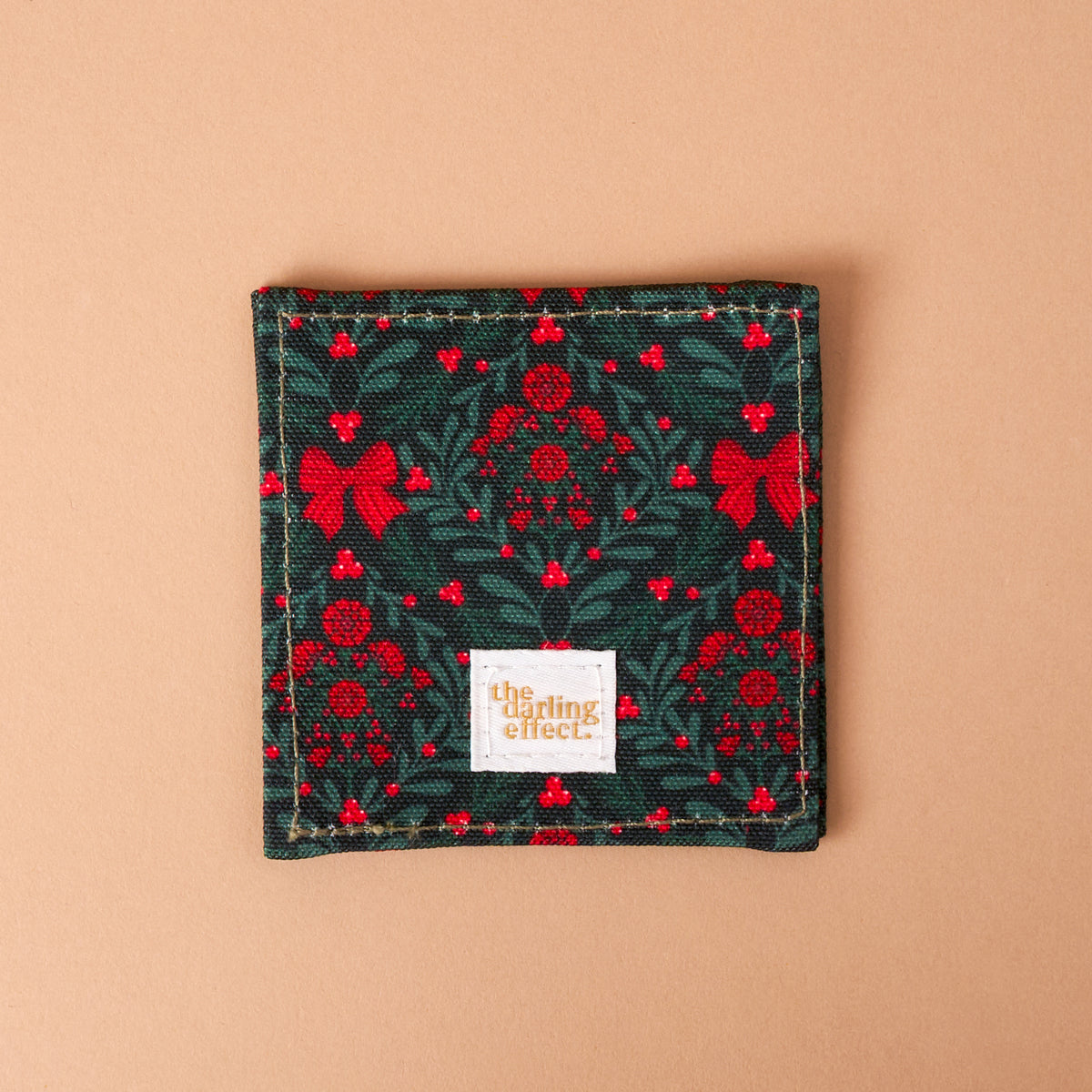 A Merry Moment Corner Keeper fabric bookmark featuring a pattern of red bows, berries, and green holly leaves, with a The Darling Effect brand tag centered at the bottom.