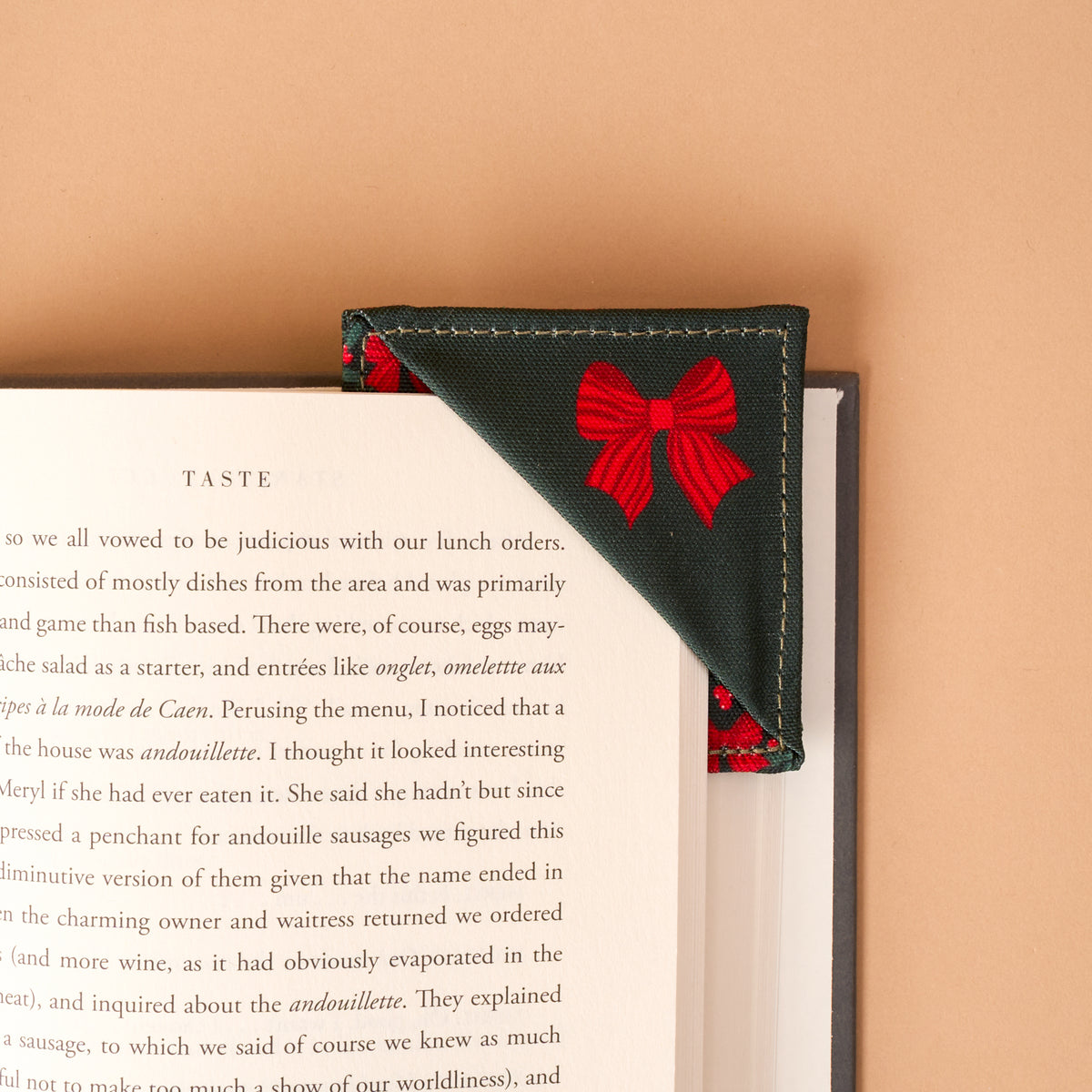 A Merry Moment Corner Keeper Fabric Bookmark in dark green with a red bow print, placed on the top corner of an open book.