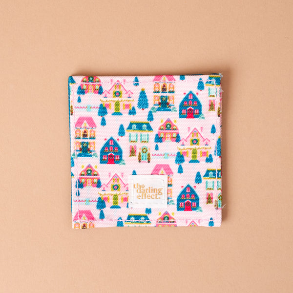 Sugarplum Lane Corner Keeper Fabric Bookmark featuring a repeating pattern of colorful houses and trees on a light pink background, with a The Darling Effect brand label sewn in the bottom corner; a perfect book lover gift.